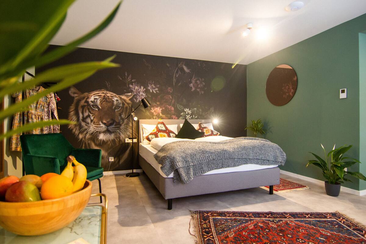 Une chambre à coucher avec un lit, un fauteuil vert, un panier de fruits et une fresque représentant un tigre sur le mur.