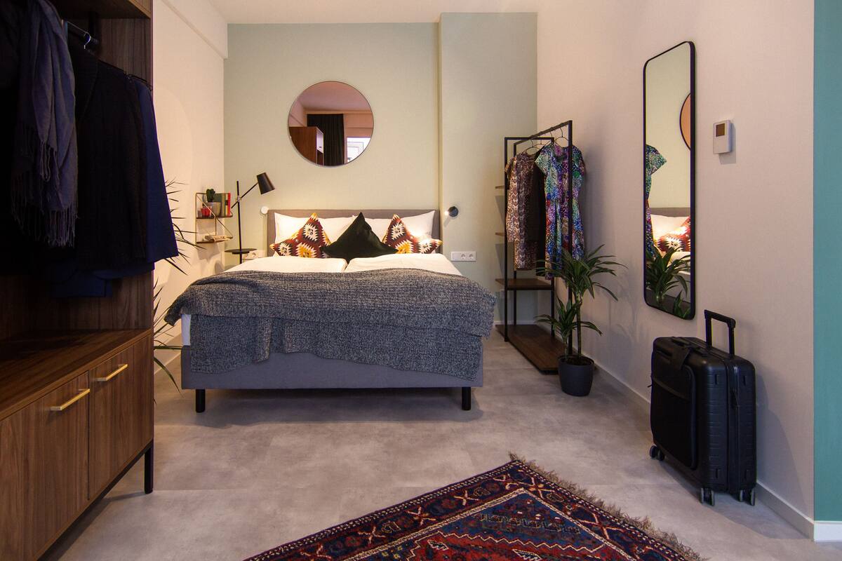 Une chambre à coucher comprenant un lit, une armoire, un miroir, un portant à vêtements, une plante en pot et une valise.