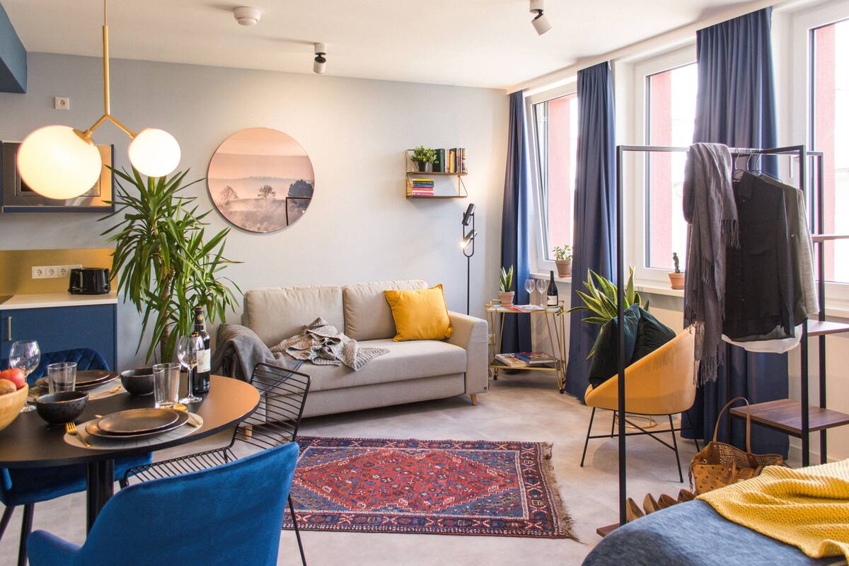 Un salon moderne avec un canapé, une table basse et un coin repas. On y trouve une grande fenêtre avec des rideaux, une plante et une bibliothèque.