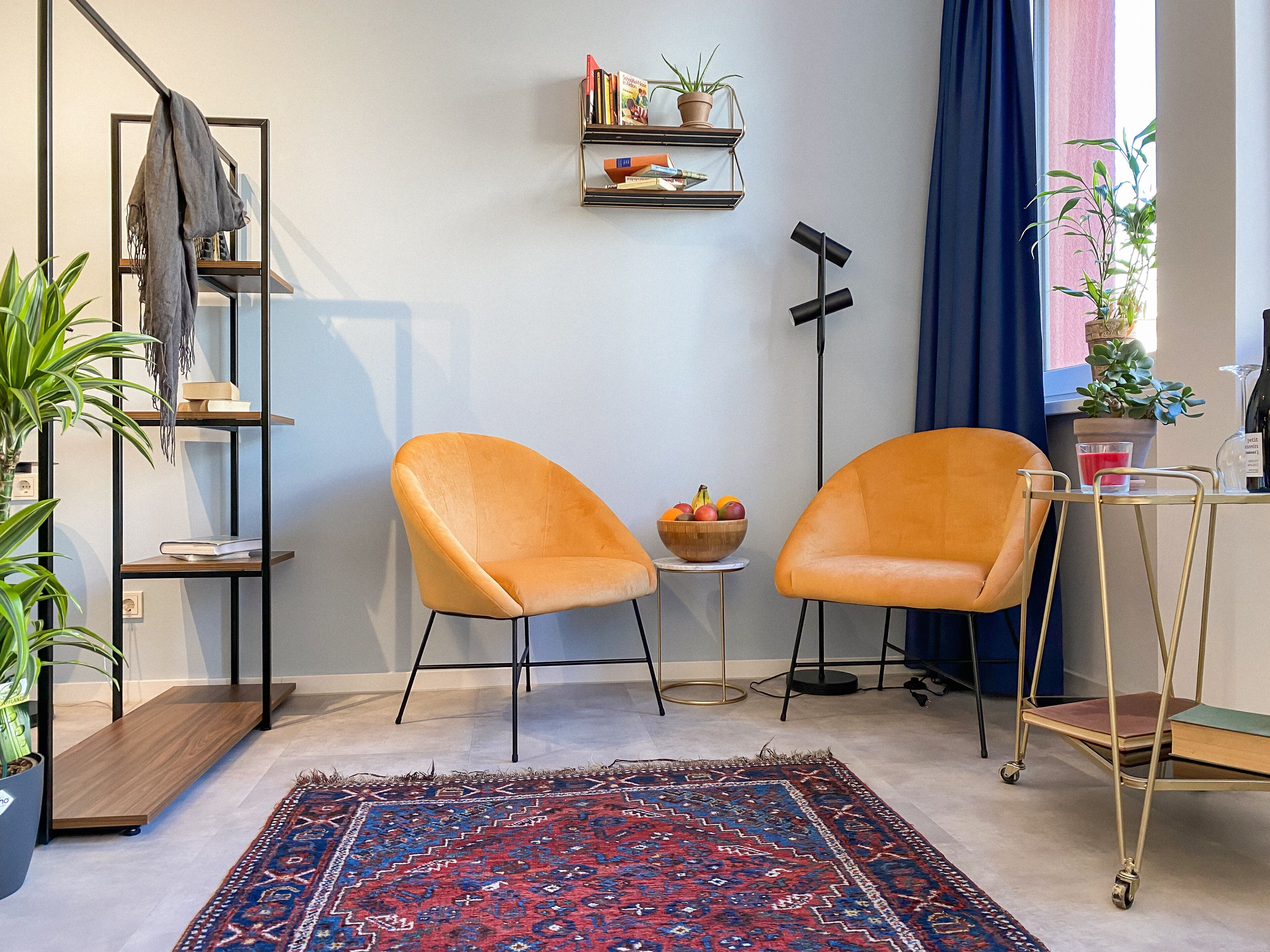 Ein modernes Wohnzimmer mit zwei orangen Sesseln, einem gemusterten Teppich, einem Bücherregal und einem kleinen Tisch mit einer Obstschale.