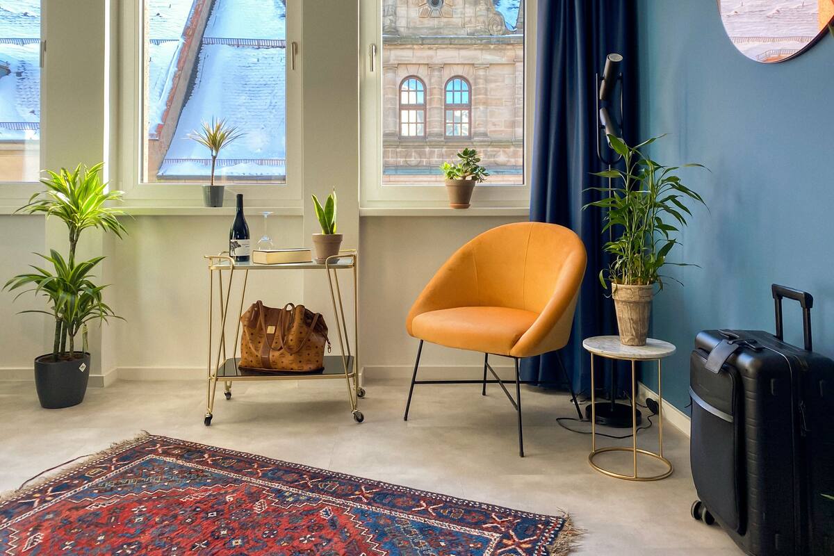 Une pièce donnant sur une rue enneigée, avec un rideau bleu, un fauteuil en cuir marron, une valise noire et un tapis à motifs.