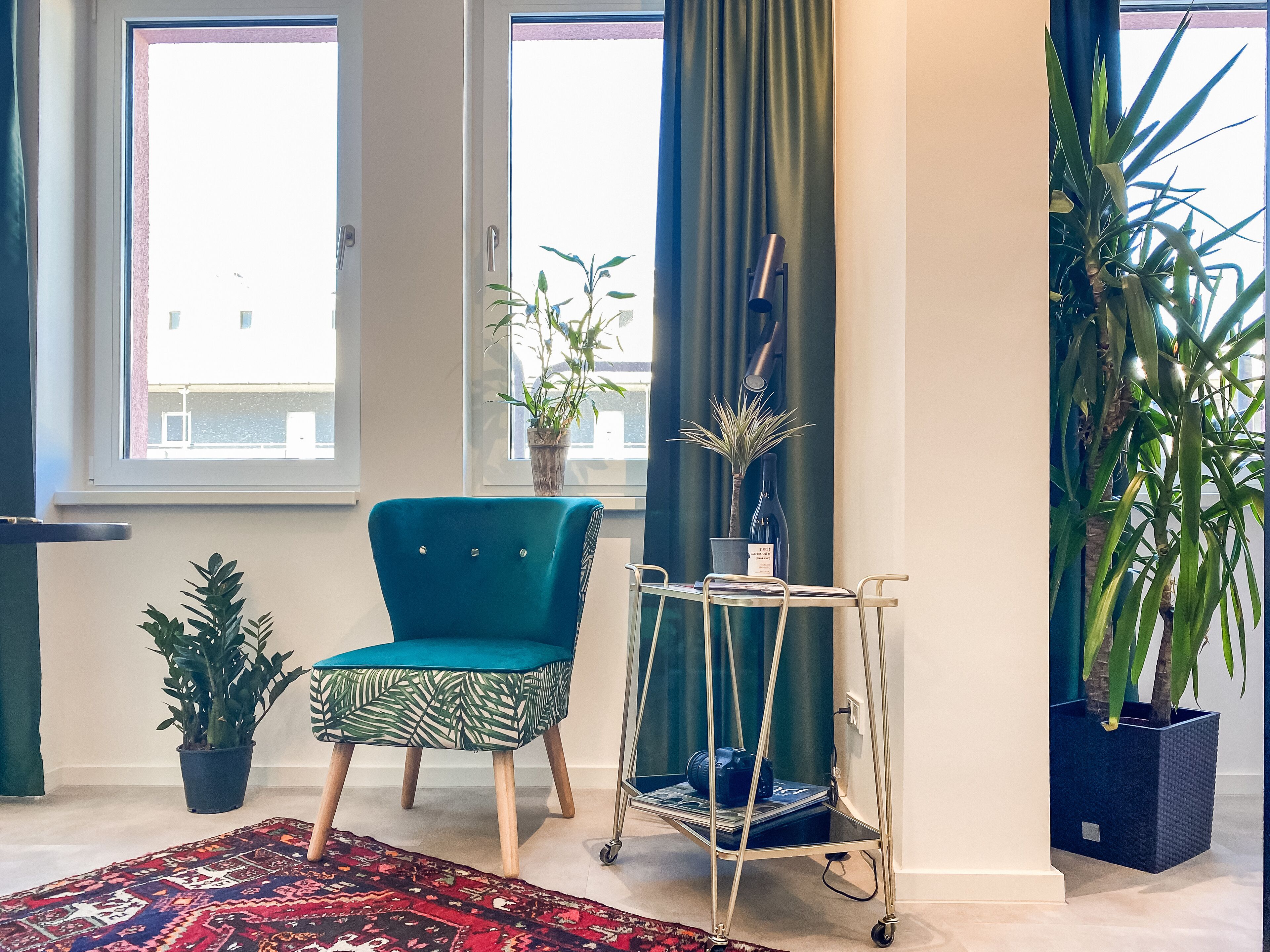 Une pièce avec un fauteuil de couleur turquoise, un pouf à motifs, une petite table sur laquelle se trouve un appareil photo, une plante en pot et un rideau foncé.