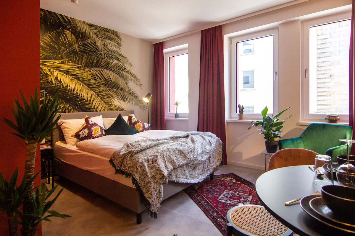 Une chambre à coucher avec un lit, une table à manger, des chaises, un rideau rouge et une fenêtre donnant sur un immeuble.