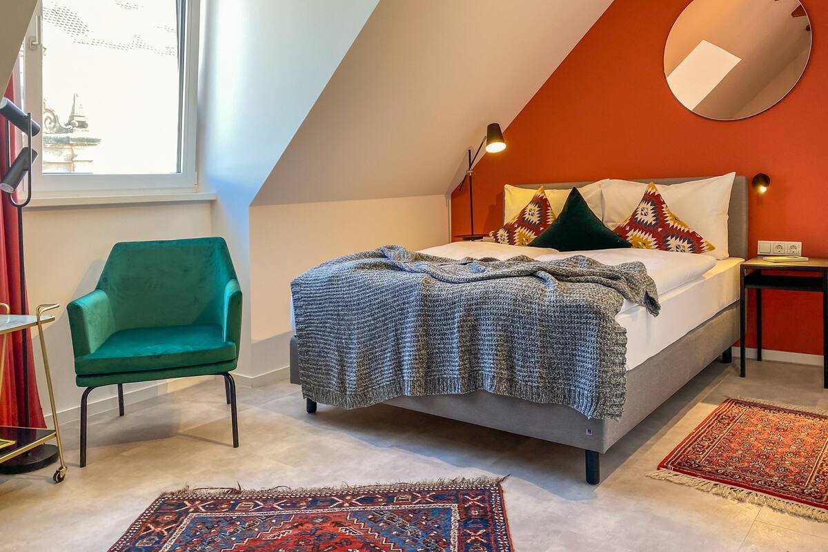 Une chambre à coucher avec un lit, un fauteuil vert, un grand miroir et une fenêtre.