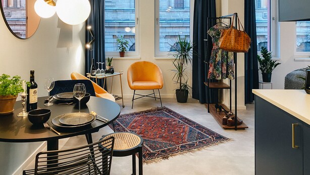 A modern apartment with a dining area, a large mirror, a potted plant, a colorful rug, and a view of the city through large windows. - Astoria Apartments (Nuremberg)