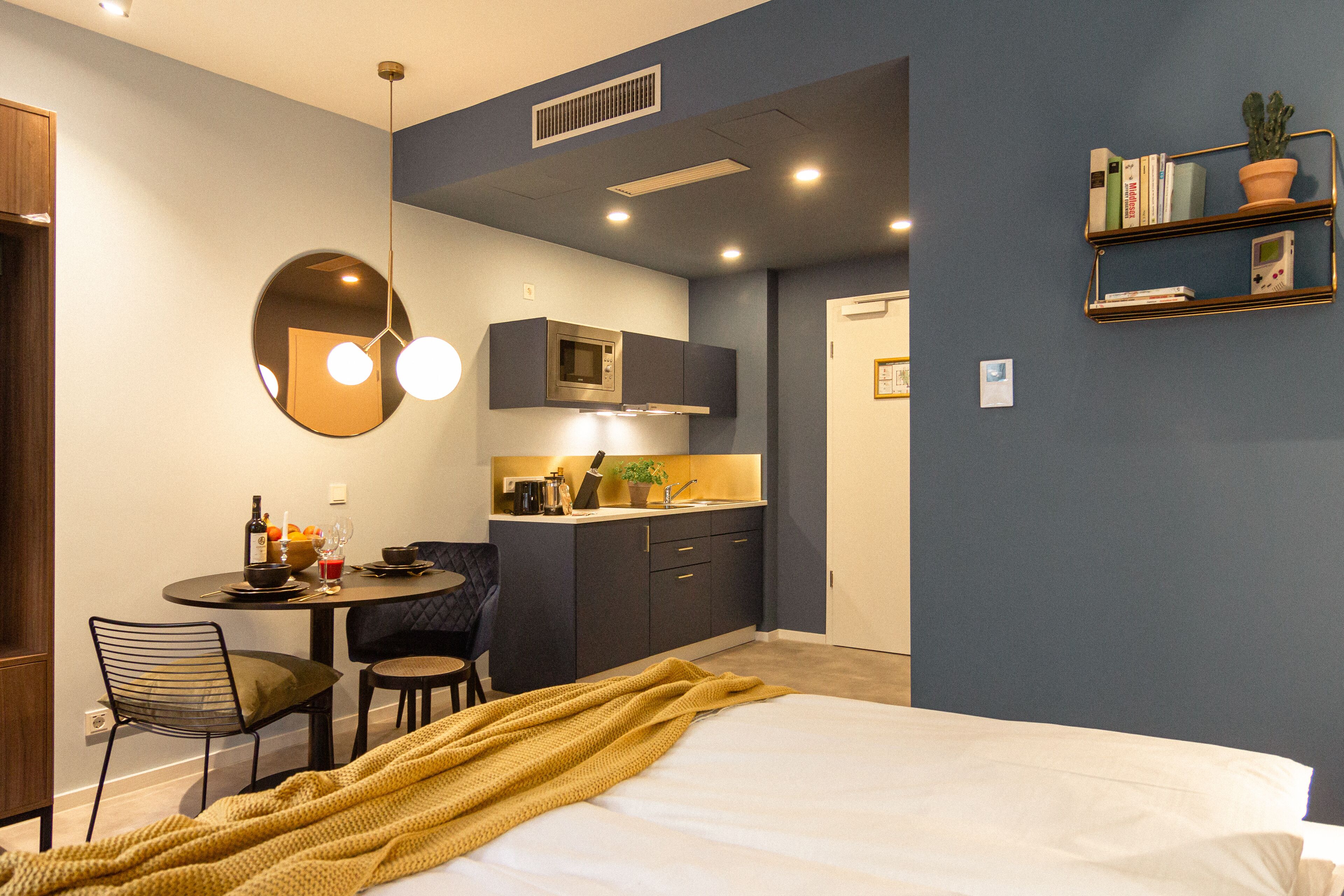 A compact hotel room with a bed, a small dining area with a table and chairs, a kitchenette, and a bookshelf.