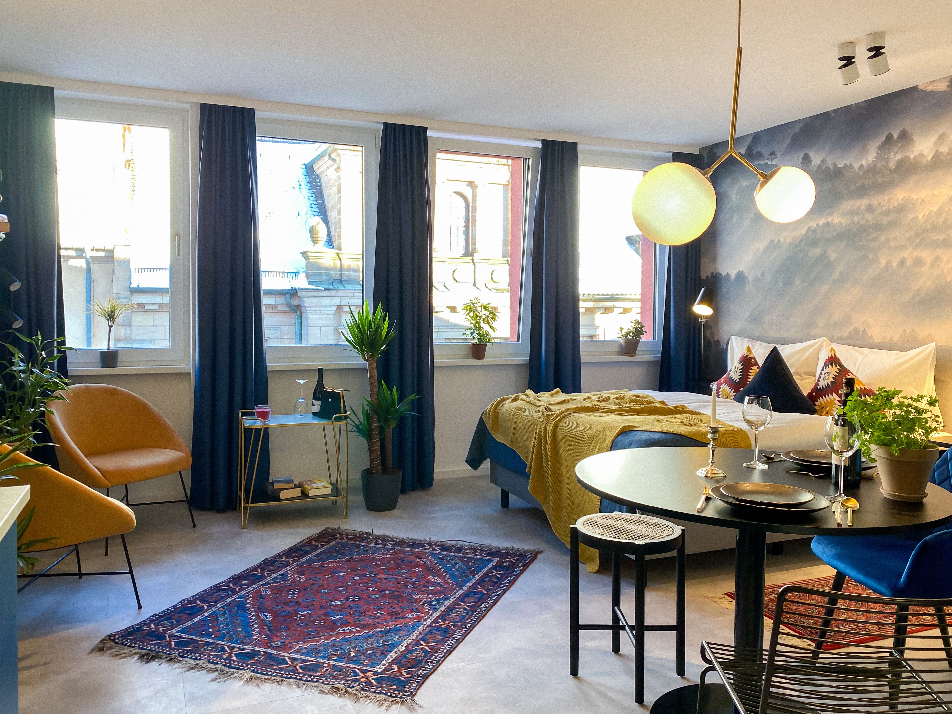 A modern hotel room with a bed, a dining table, a sofa, and a view of the city through large windows.