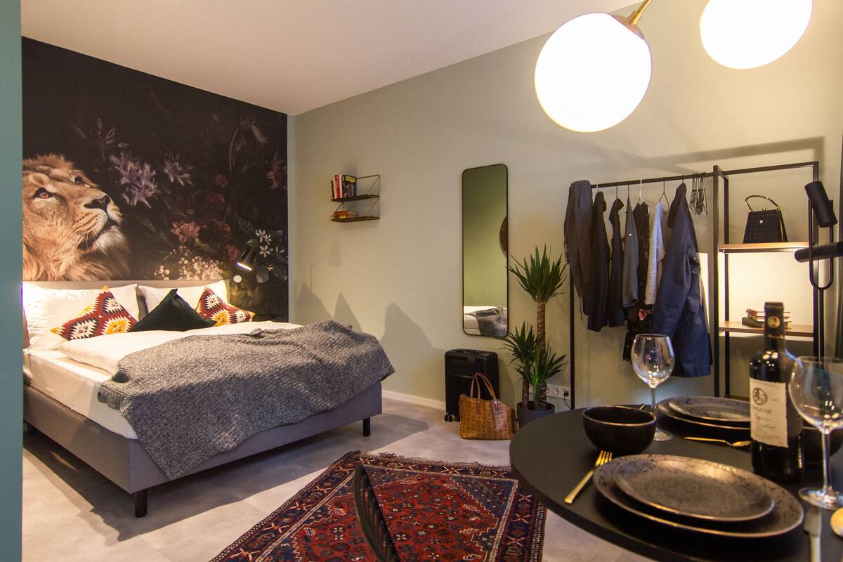 Une chambre moderne avec un grand lit, un miroir, une armoire et un coin repas avec une table dressée pour deux personnes, ainsi qu’un mur orné d’une fresque florale.