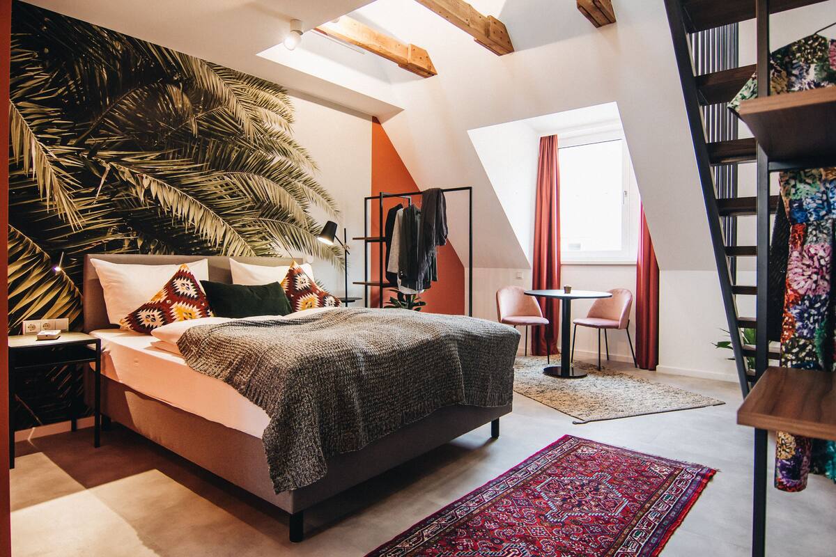 Une chambre à coucher comprenant un lit, une table de chevet, une chaise, une petite table, une bibliothèque et une grande fenêtre.