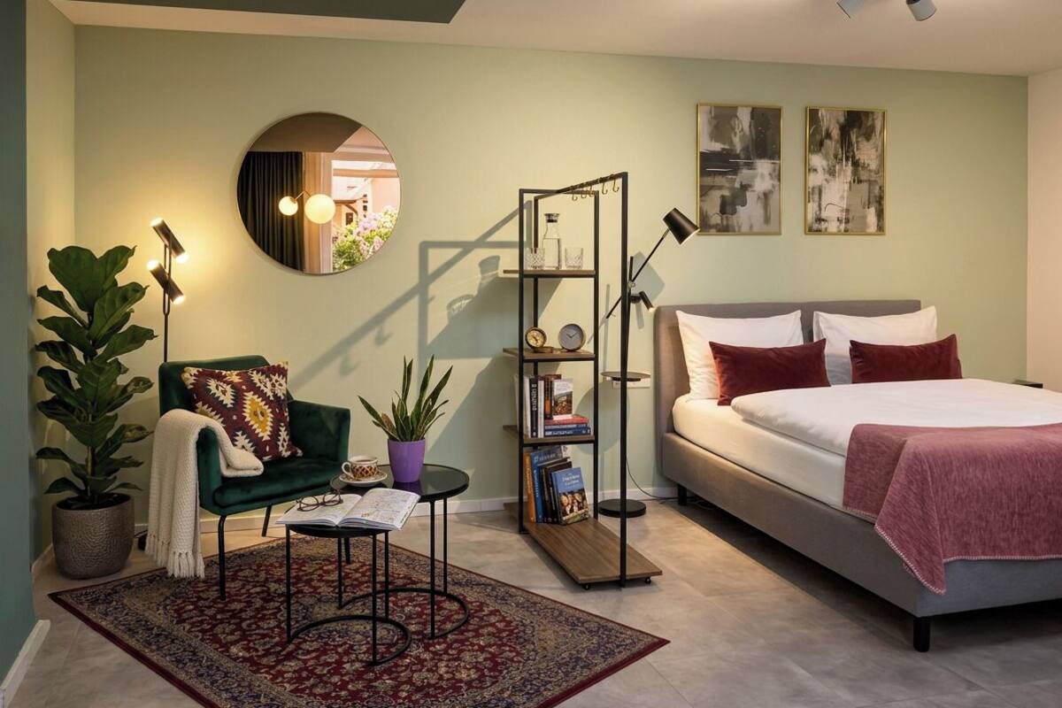 Une chambre à coucher comprenant un lit, une chaise, une petite table, une bibliothèque, un miroir et une plante en pot.