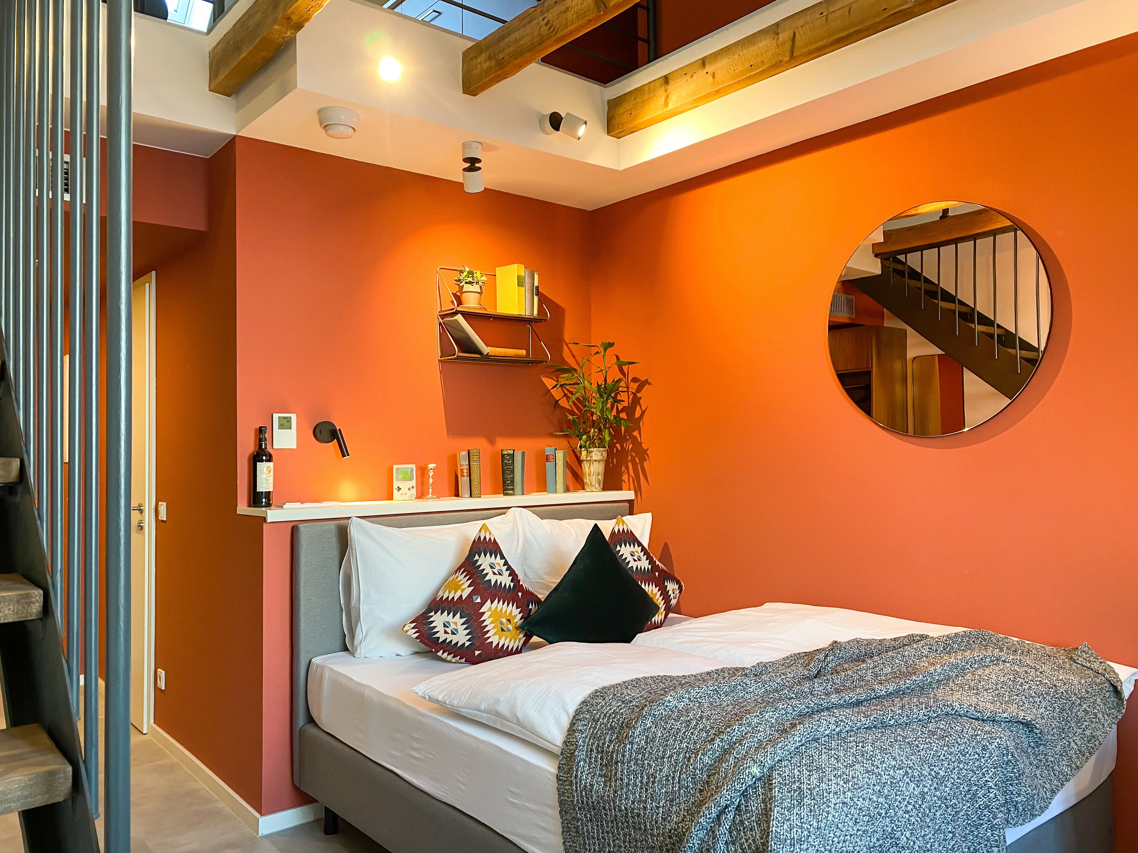 A bedroom with a bed, a round mirror, a shelf with books and a plant, and a staircase.