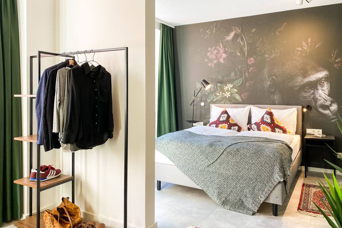 Une chambre avec un lit, une étagère fixée au mur où sont rangés des vêtements et des chaussures, une table de chevet avec une lampe, et une grande fresque florale sur le mur.