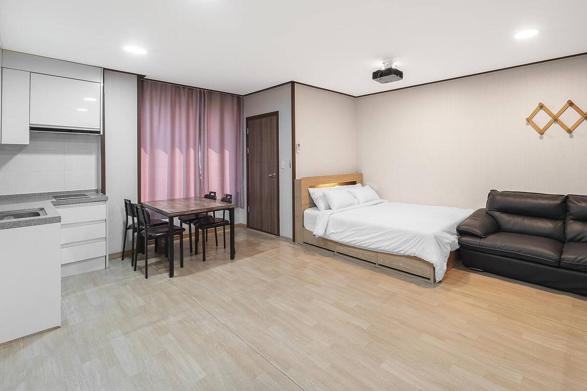 A compact hotel room with a bed, a sofa, a dining table with chairs, and a kitchenette.