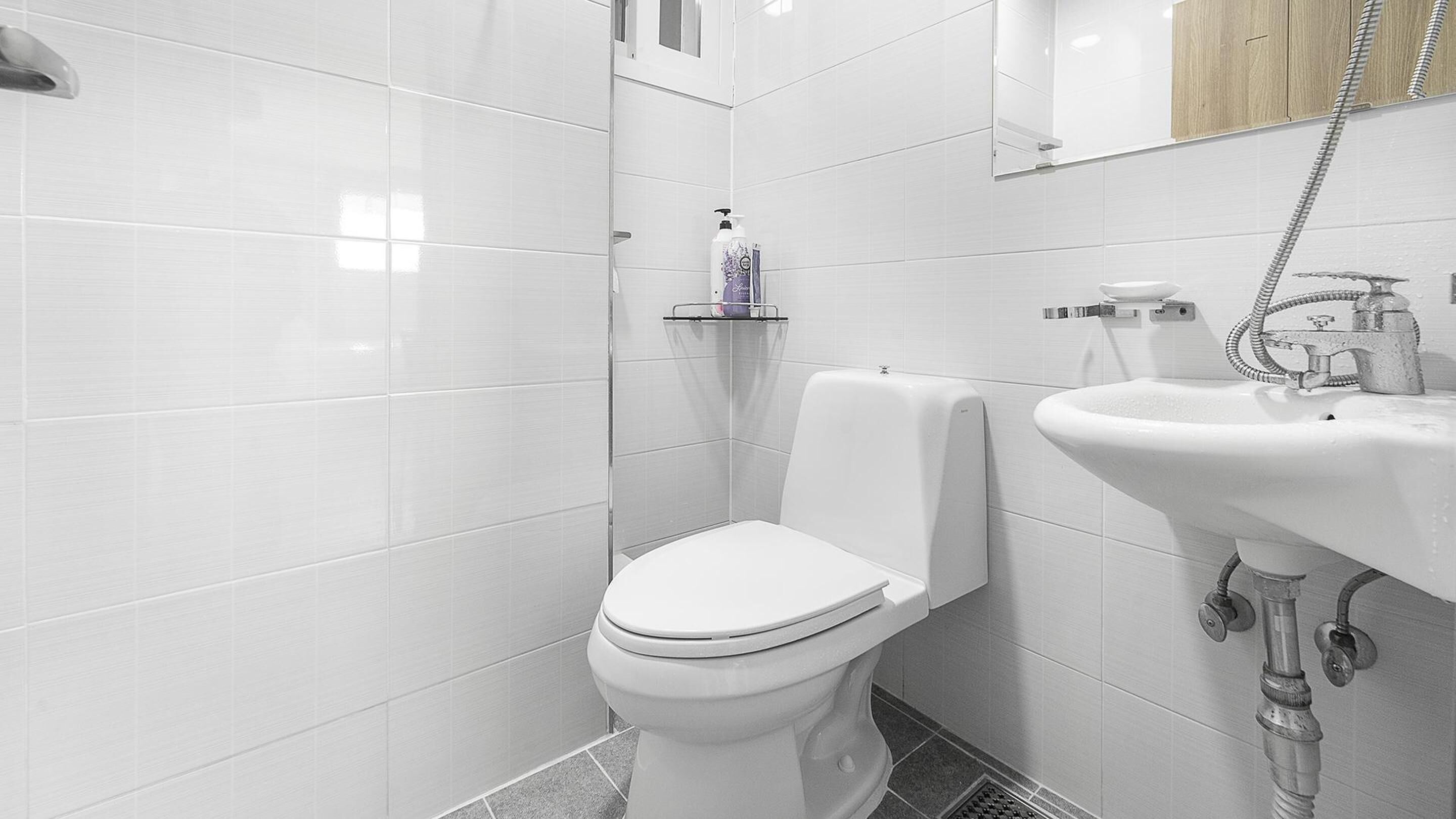 Et moderne badeværelse med hvide fliser, toilet, håndvask og bruser.