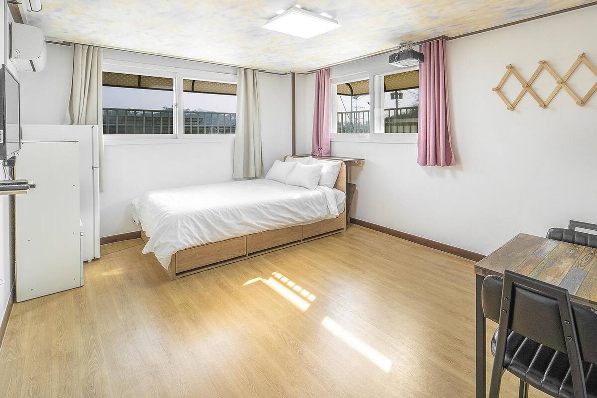 A small, well-lit room with a single bed, a wooden table, and a chair. There are two windows with curtains, a ceiling light, and a wall-mounted projector.