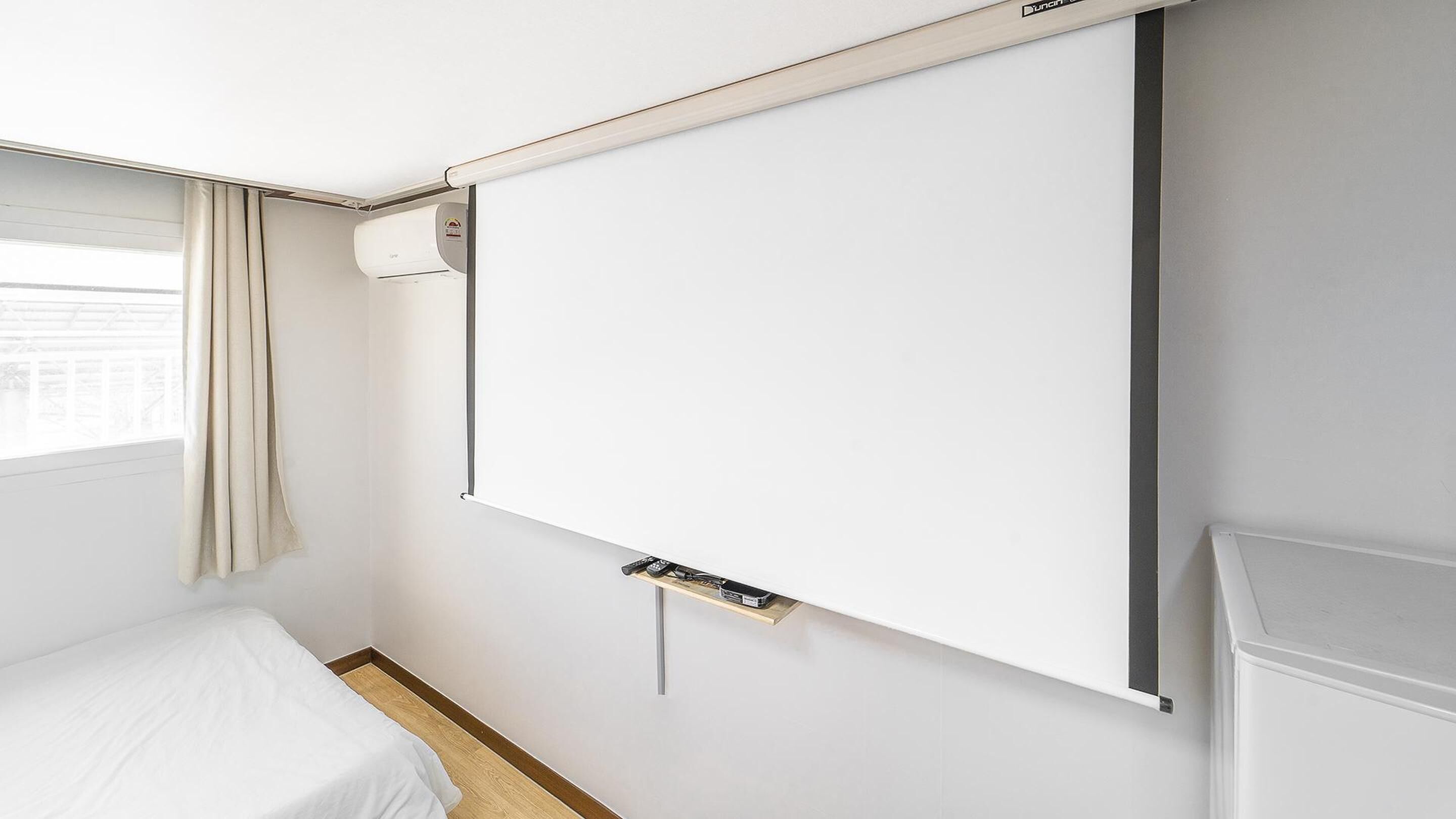 Une chambre d’hôtel avec un grand téléviseur blanc fixé au mur, un lit avec une literie blanche, un plancher en bois et une fenêtre avec des rideaux.