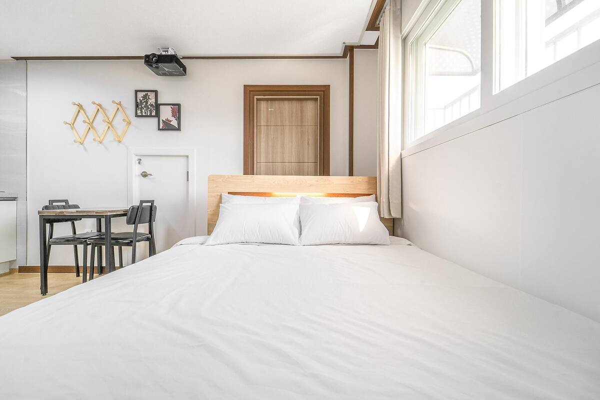 A bedroom with a bed, a wooden headboard, a desk with chairs, a door, and a window.