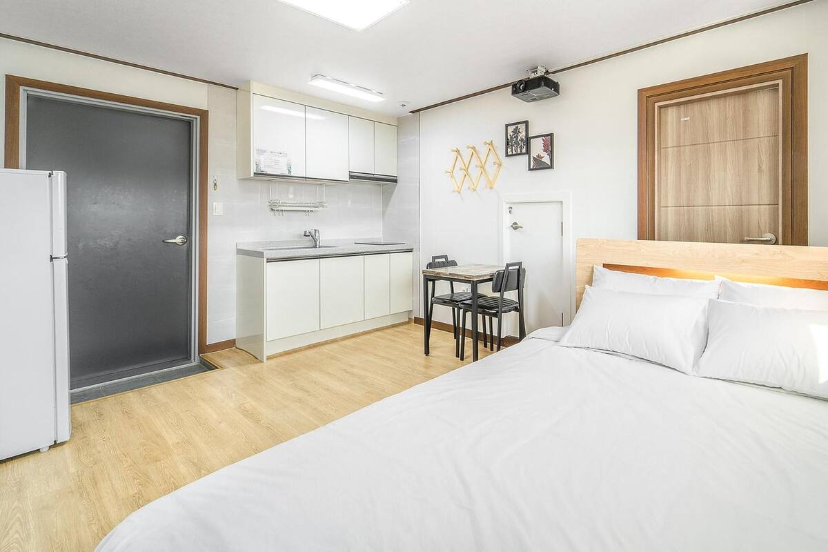 A hotel room with a bed, a desk, a chair, a small kitchenette, and a door.