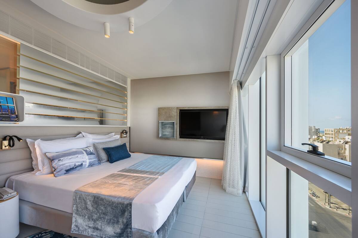 A modern hotel room with a large bed, a flat-screen TV mounted on the wall, and a view of the city through large windows.