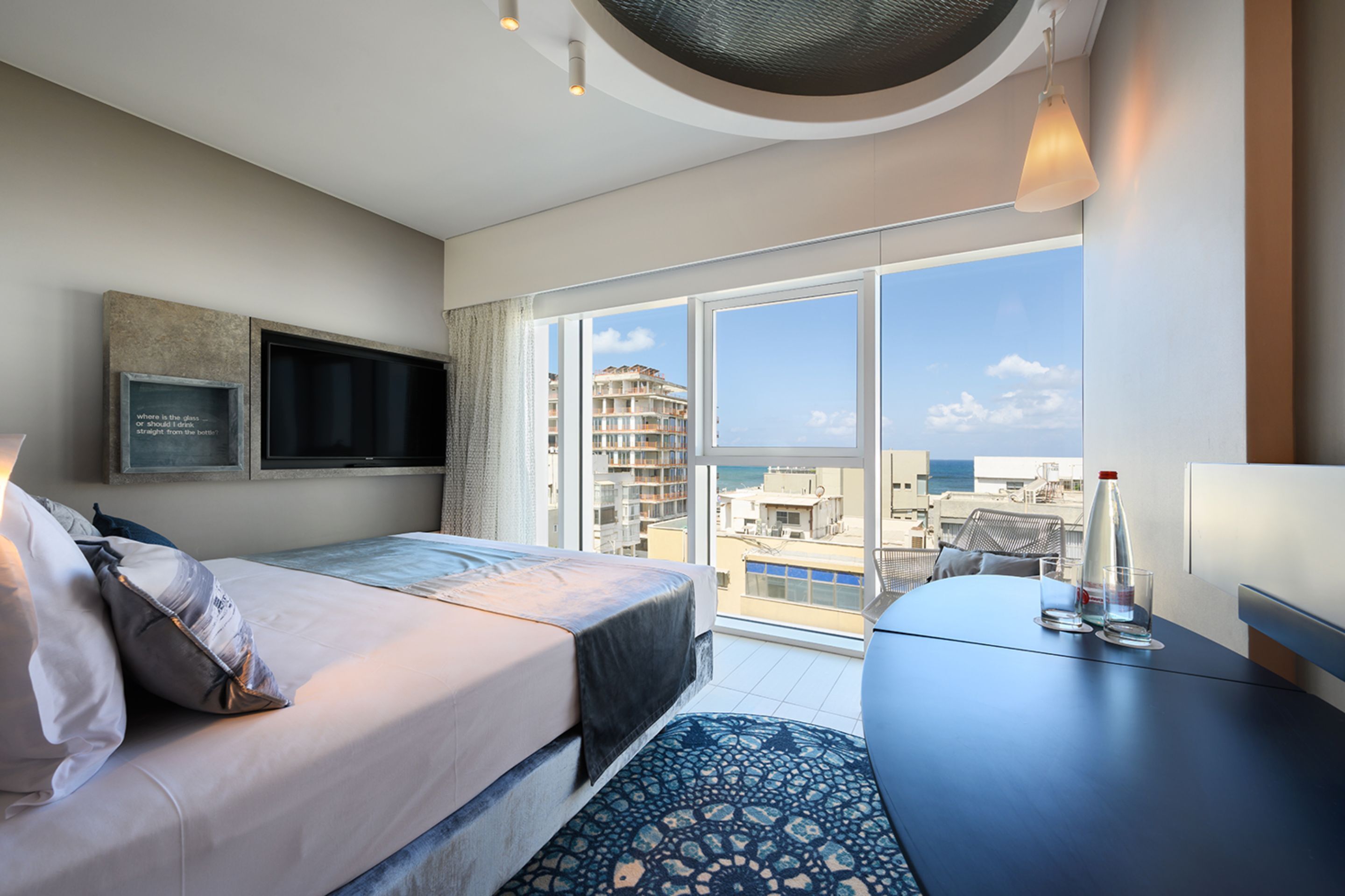 Una habitación de hotel moderna con una cama grande, un televisor de pantalla plana en la pared, una mesa de comedor con una botella y vasos, y vistas a la ciudad y al mar a través de grandes ventanales.