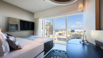 Una habitación de hotel moderna con una cama grande, un televisor de pantalla plana en la pared, una mesa de comedor con una botella y vasos, y vistas a la ciudad y al mar a través de grandes ventanales.
