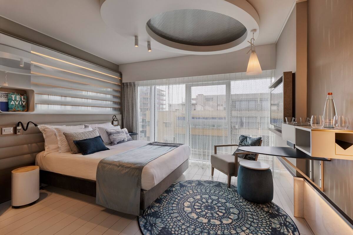 A modern hotel room with a large bed, a desk, and a view of the city through large windows.