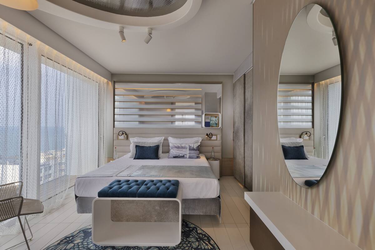 A modern bedroom with a large bed, bedside tables, a mirror, and a window with blinds.