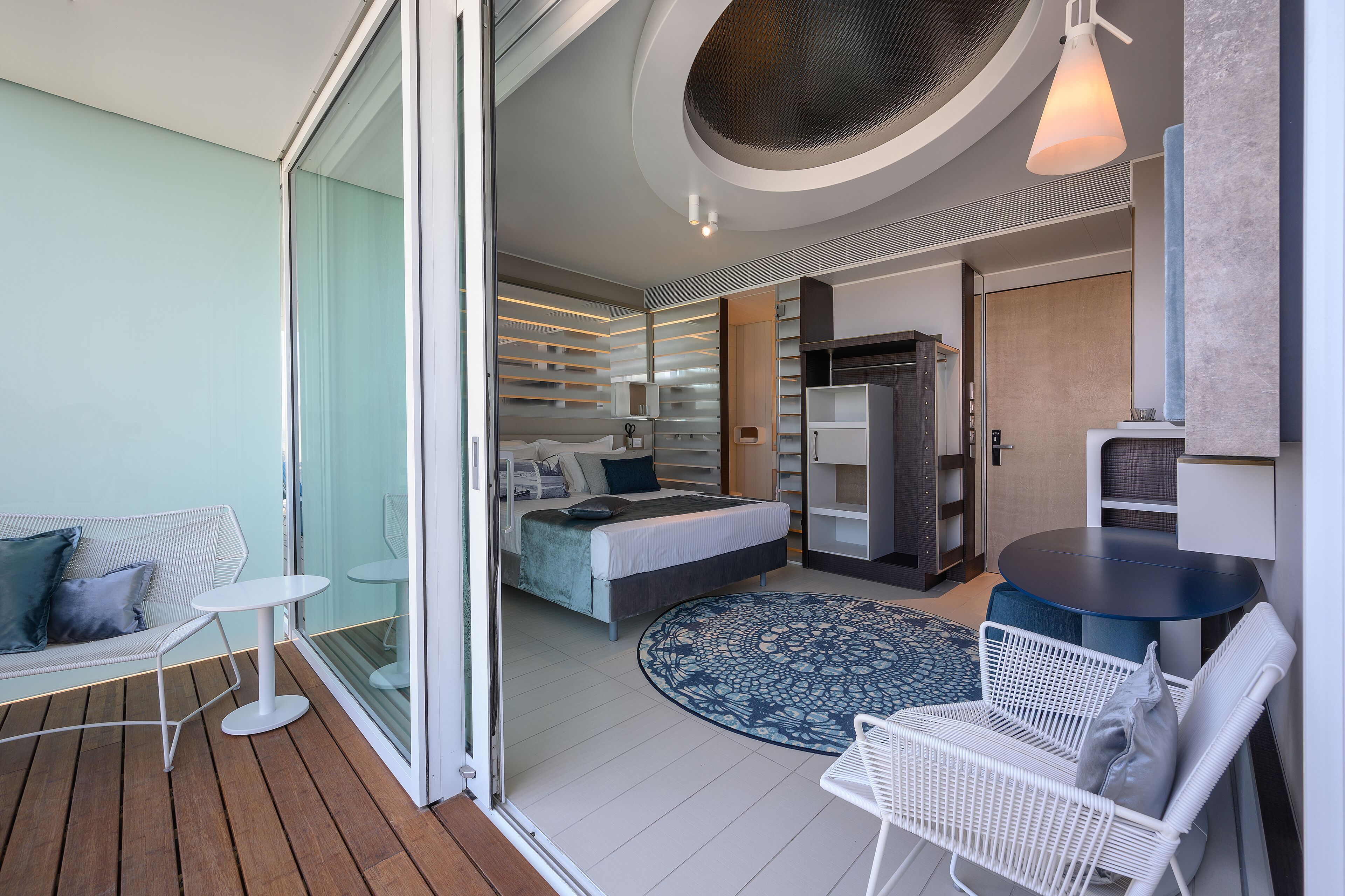 Une chambre d’hôtel moderne avec un lit, une petite table ronde, une chaise blanche, un canapé et une grande fenêtre.