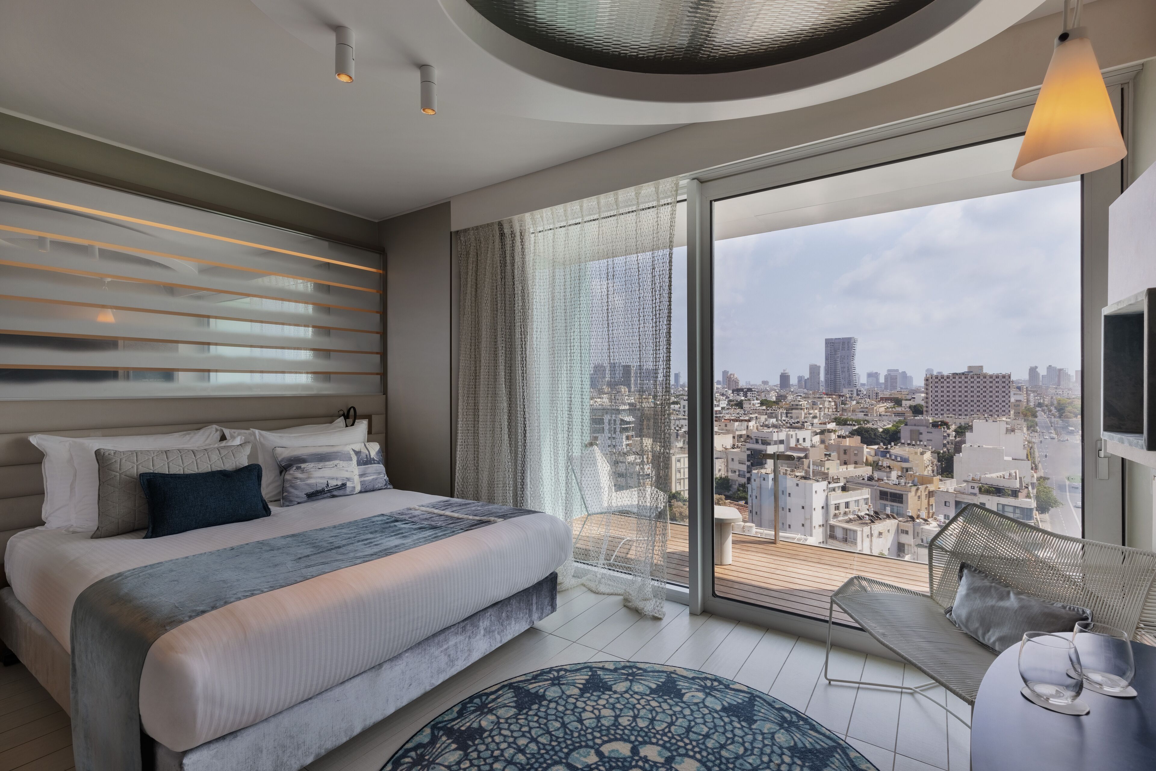 Camera da letto moderna con un letto grande, vista sulla città e un balcone con una sedia e un tavolo.