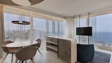 Una sala de estar moderna con una mesa de comedor redonda, sillas color beige, un reposapiés azul oscuro, un televisor de pantalla plana y vistas de la ciudad a través de amplias ventanas.