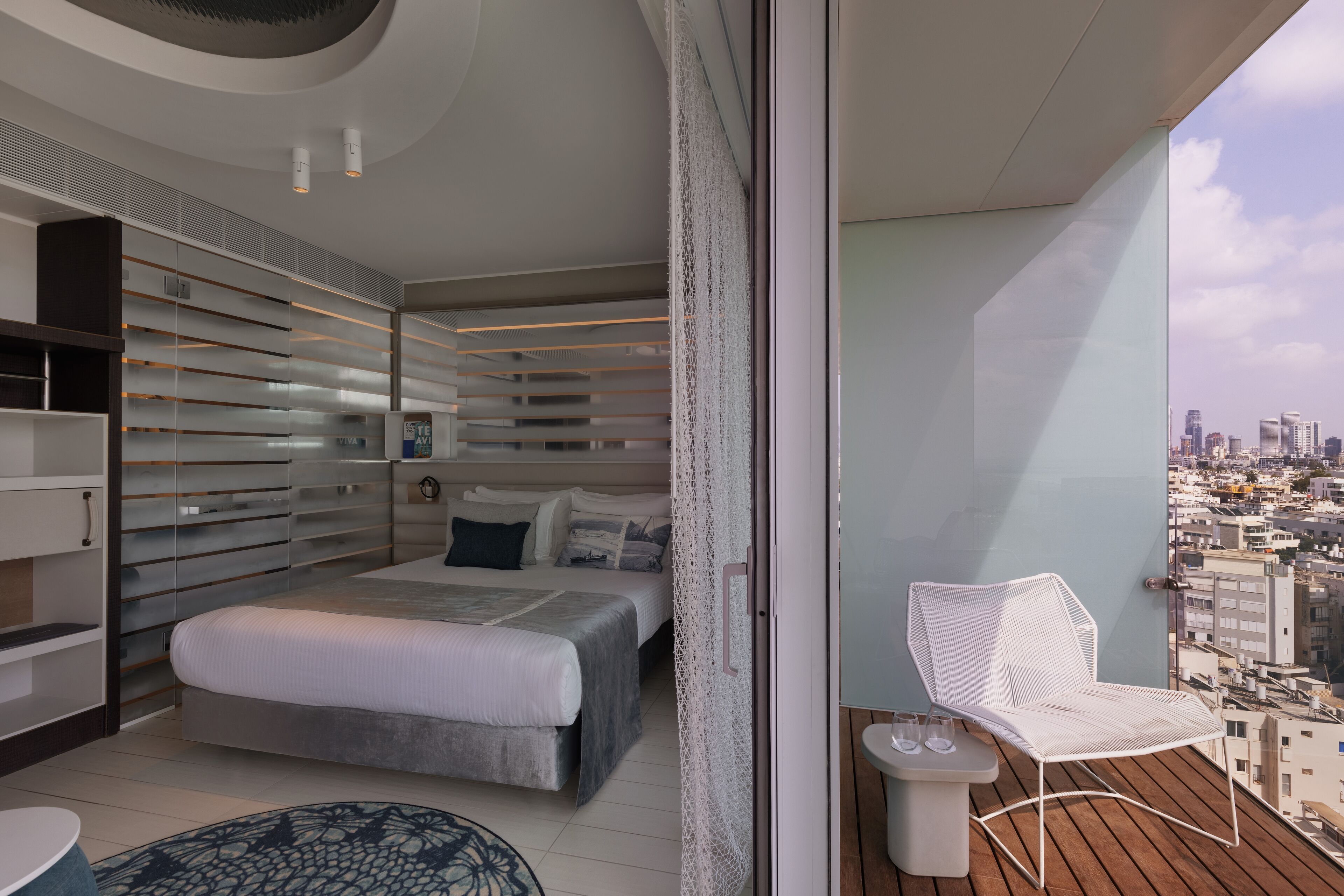 Une chambre moderne avec un grand lit, un balcon avec une chaise blanche et une vue sur la ville.
