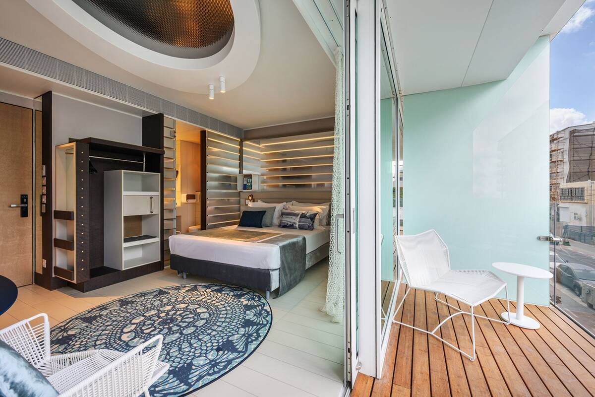 A modern hotel room with a bed, a seating area with a table, and a balcony with a view of the city.