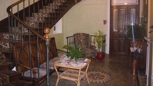 A staircase with a wooden handrail and a patterned carpet. There is a chair with a floral pattern and a small table with a plant. A door with a decorative frame is visible in the background. -  Sarl Aux Studios Du Parc (Le Val-d'Ajol)