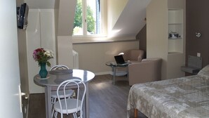 A compact room with a bed, a small table with a laptop, a chair, a sofa, and a window providing natural light. -  Sarl Aux Studios Du Parc (Le Val-d'Ajol)