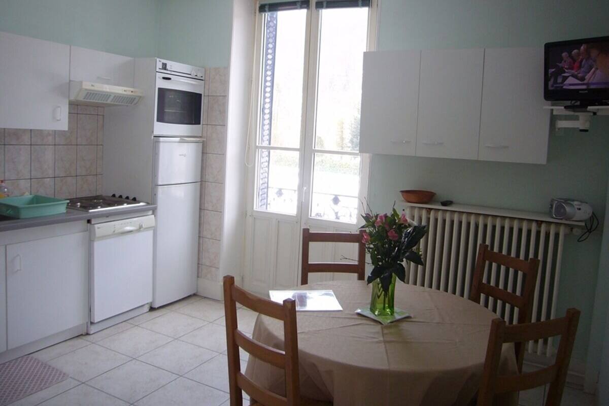 Una cocina con armarios blancos, una estufa, un refrigerador, una mesa de comedor con sillas y una ventana con cortinas.