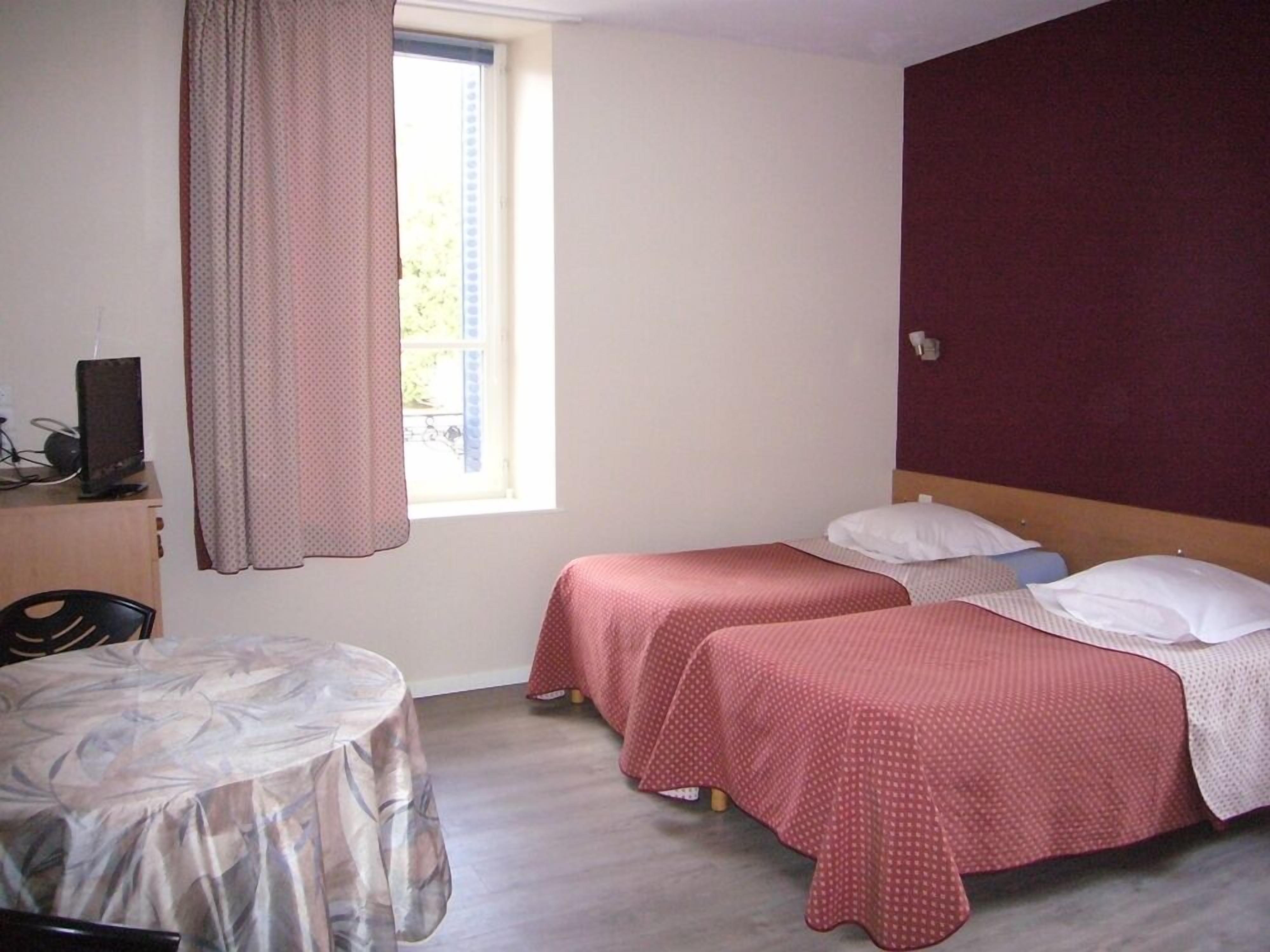 A small, simple room with two beds, a desk with a computer, and a window with curtains.
