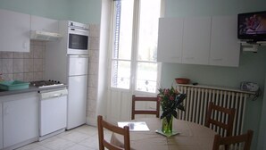 A kitchen with white cabinets, a stove, a refrigerator, and a dining table with chairs. There is a window with curtains and a radiator above the table. -  Sarl Aux Studios Du Parc (Le Val-d'Ajol)