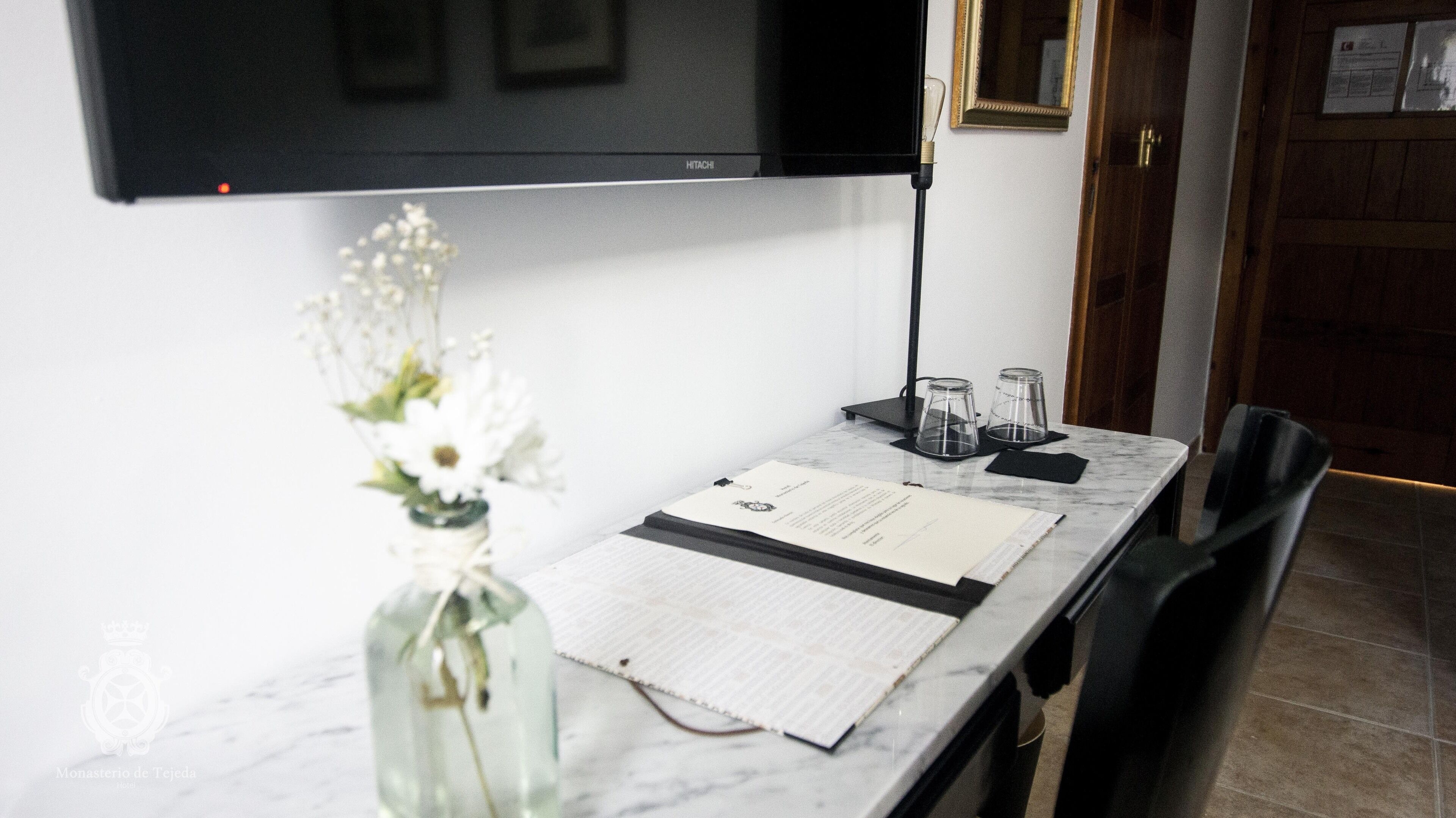A hotel room with a flat-screen TV mounted on the wall, a small table with a menu, a vase with flowers, and a glass carafe.