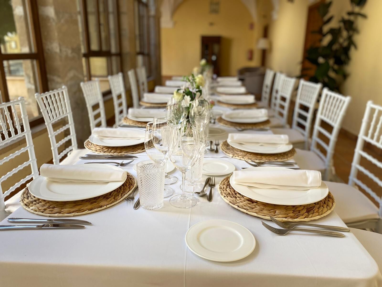 A long dining table set with white plates, silverware, and clear glassware, surrounded by white chairs with woven placemats.