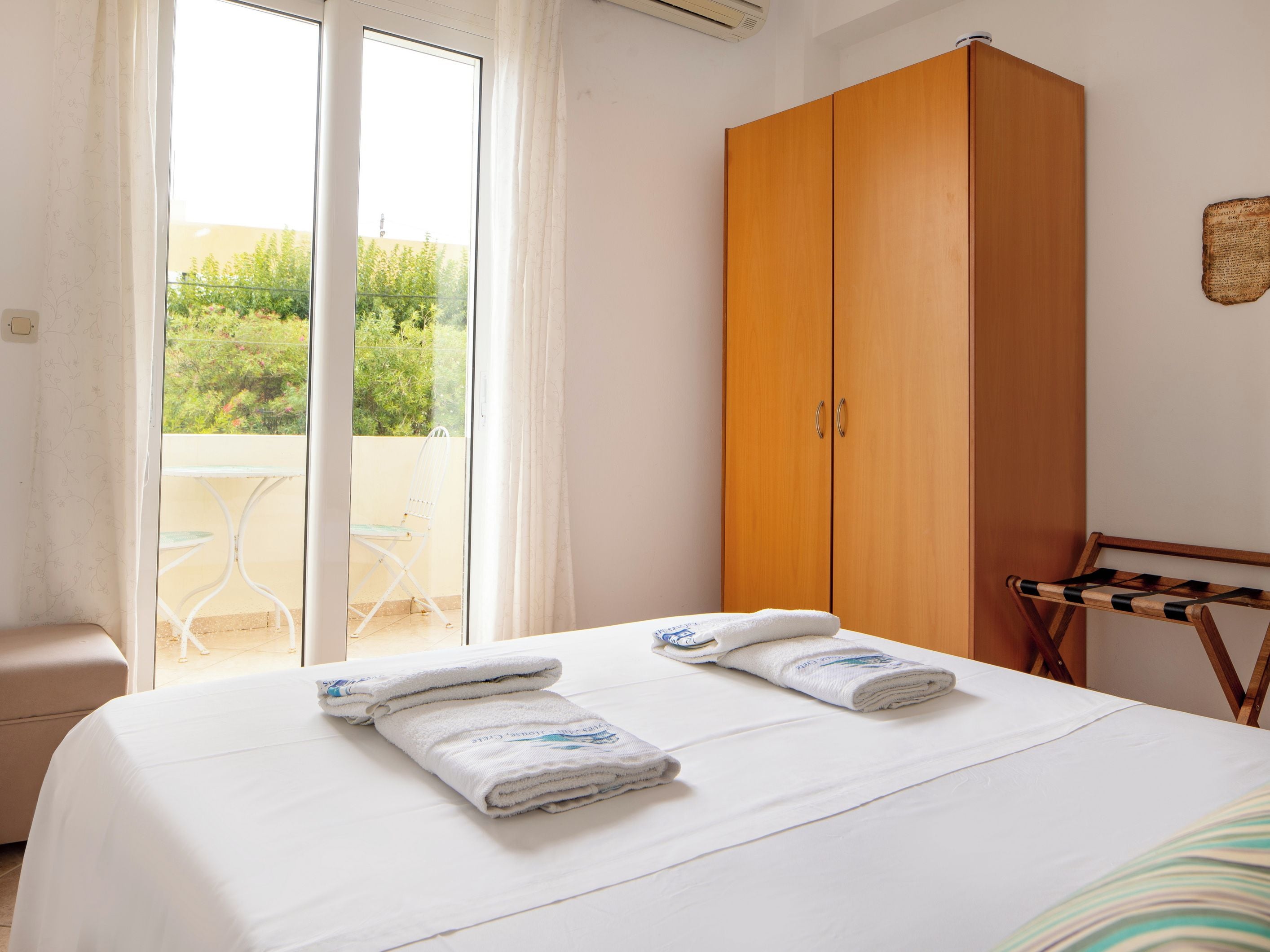 A bedroom with a bed, two towels, a wooden wardrobe, a balcony with a chair, and a window with curtains.