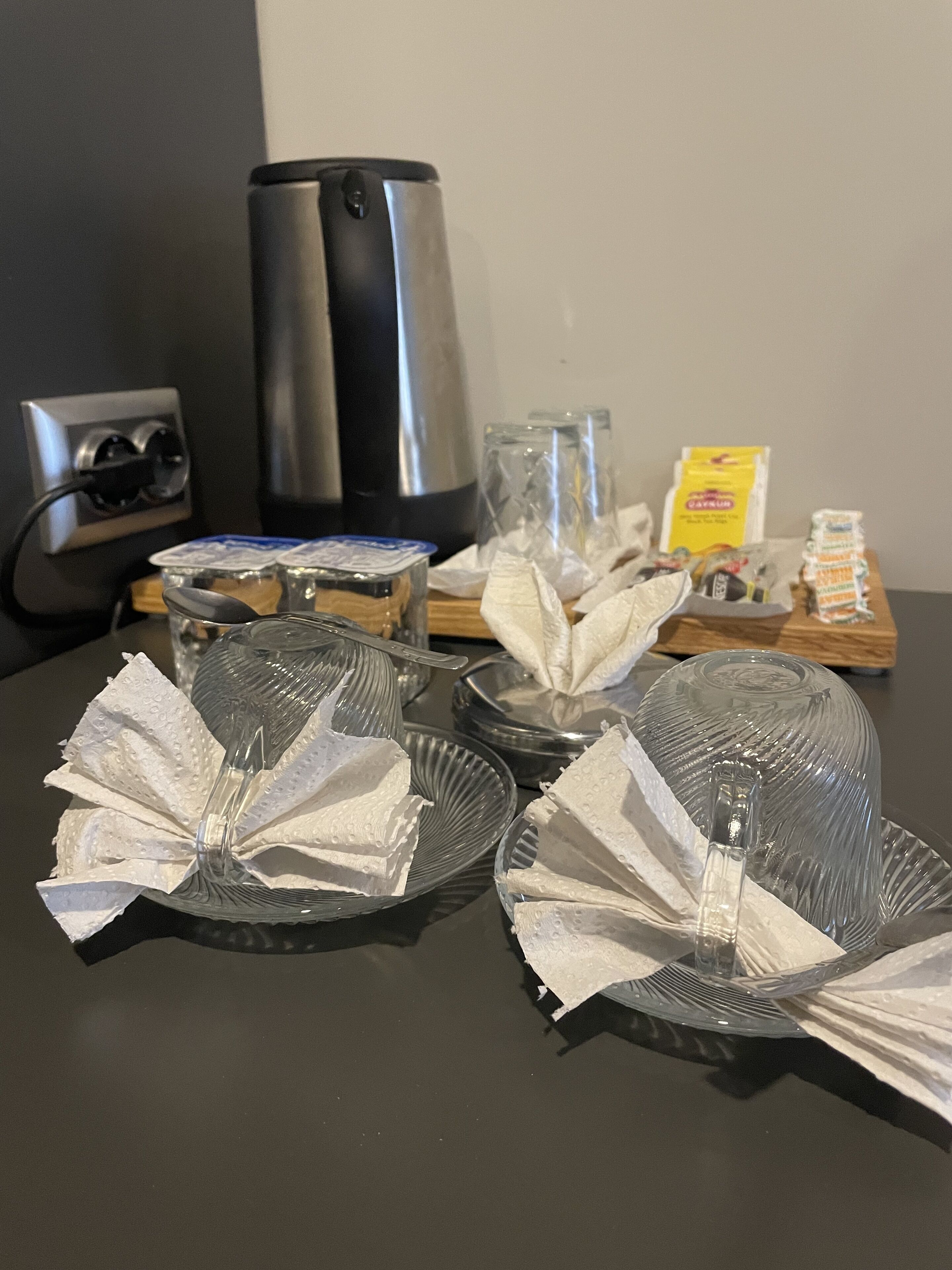 Une table avec de la vaisselle, des serviettes et des condiments, une machine à café et une prise électrique.