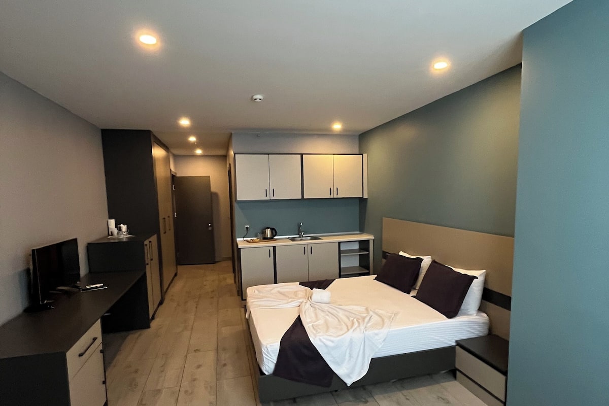 A modern hotel room with a bed, bedside tables, a desk, and a kitchenette.