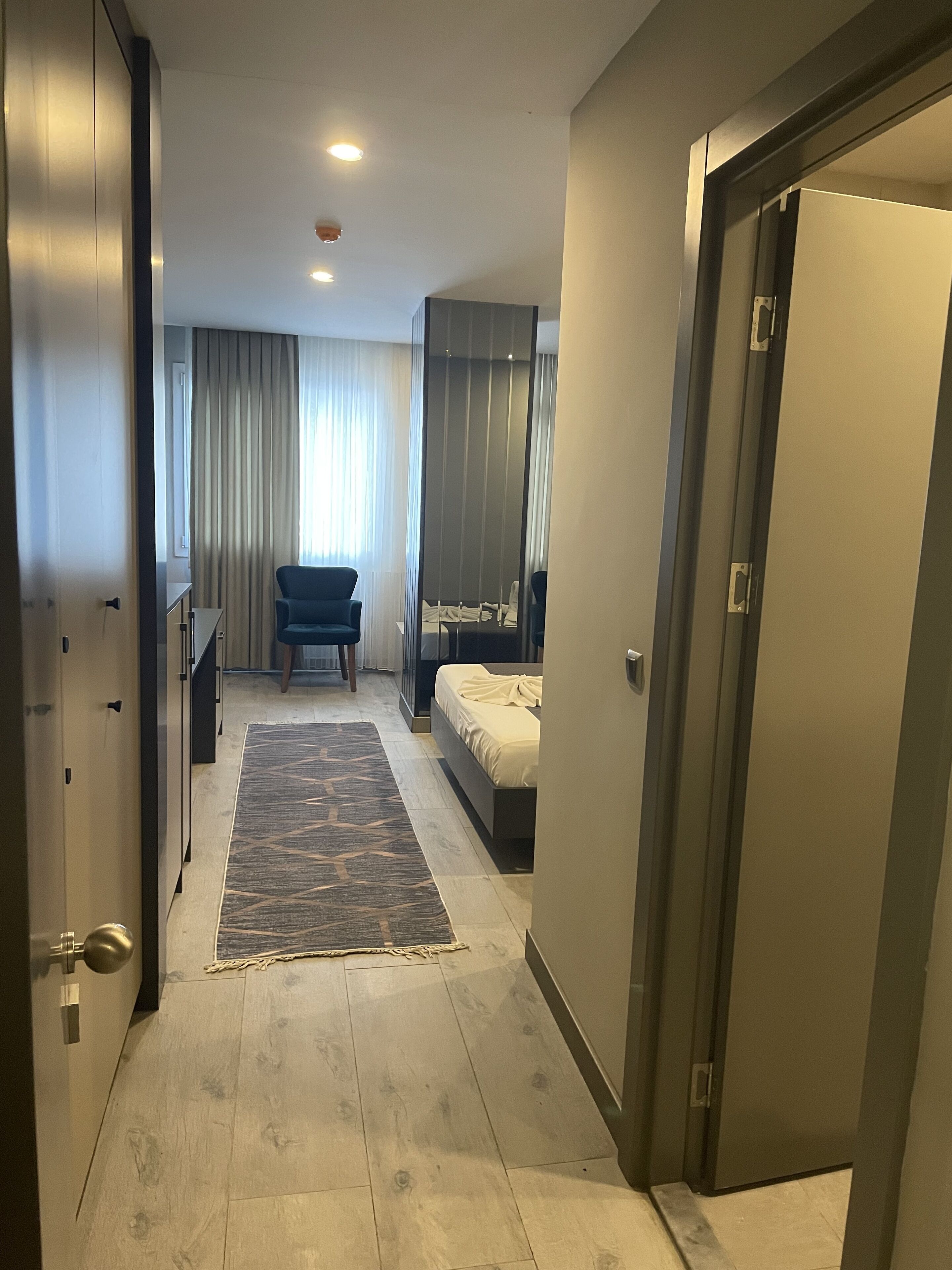 A modern hotel room with a bed, a chair, a desk, and a wardrobe.