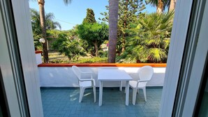 A balcony with a white table and chairs, overlooking a garden with various trees and plants.