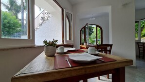 A dining area with a wooden table, chairs, a potted plant, and a view of greenery outside.