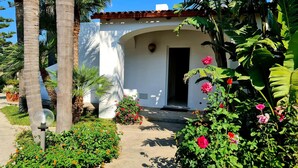 A white building with a red-tiled roof, surrounded by lush greenery and flowering plants.