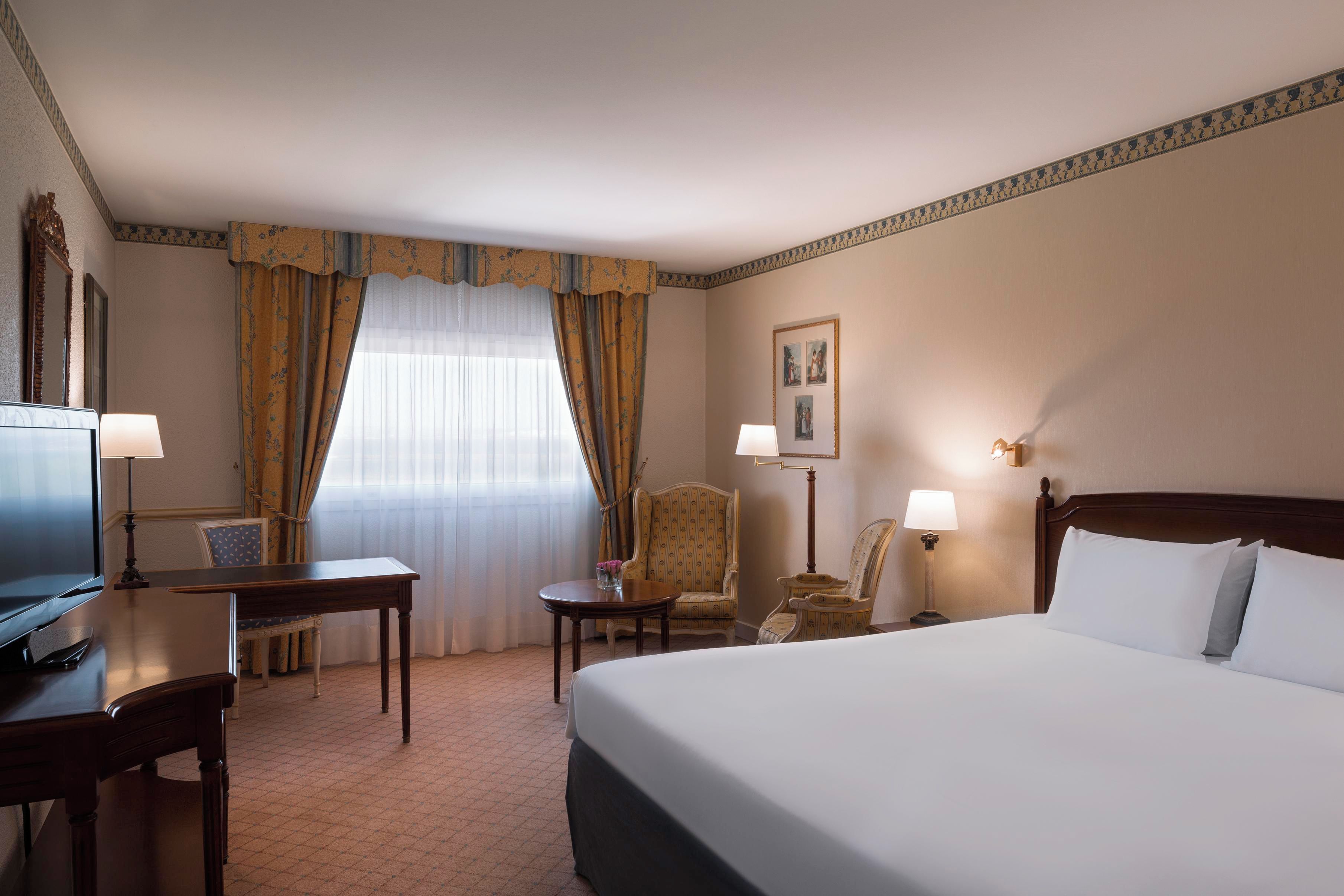 A hotel room with a bed, a desk, a chair, a television, and a window with curtains.