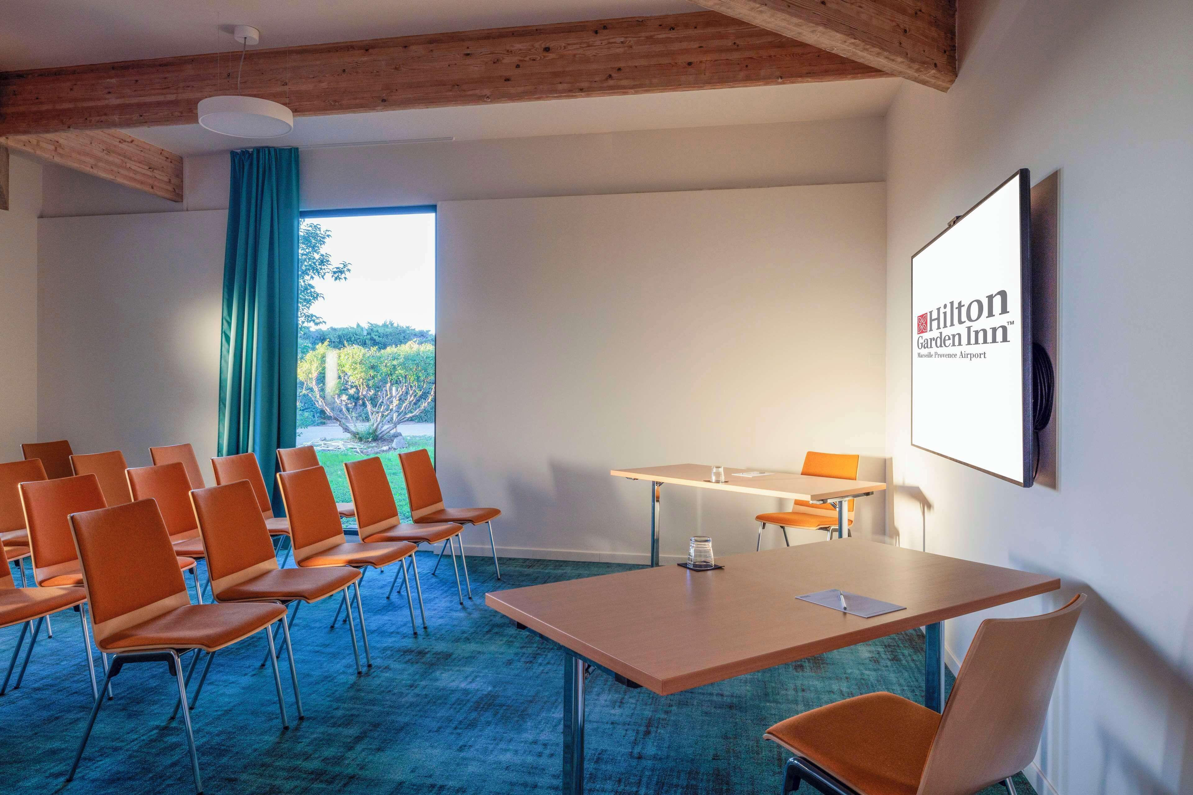 A conference room with a large screen displaying Hilton Garden Inn logo, a wooden ceiling, and a view of greenery outside.