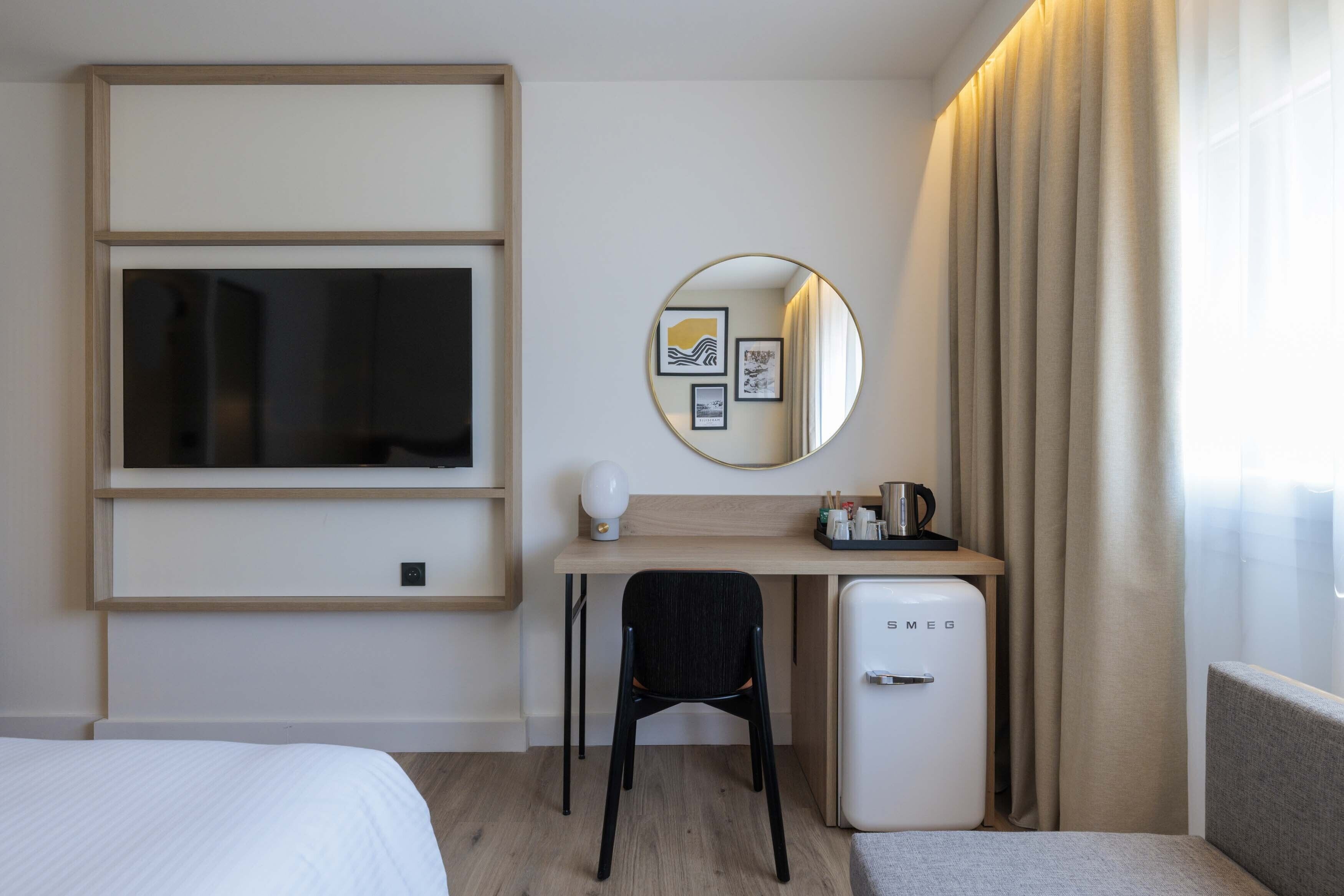 Une chambre d’hôtel comprenant un lit, un bureau avec une chaise, un petit réfrigérateur, un miroir et une fenêtre avec des rideaux.