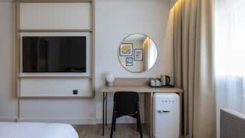 A hotel room with a bed, a desk with a chair, a small refrigerator, a mirror, and a window with curtains.
